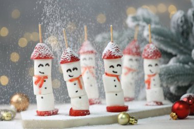 Masadaki arka planda bulanık ışıklarla marşmelovlardan, çileklerden ve Noel dekorundan yapılmış komik kardan adamların üzerine pudra şekeri serpmek, yakın plan. Bokeh etkisi