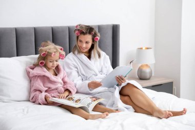 Anne ve kız bornozlu ve saç silindirli evde yatakta dergi okuyor.