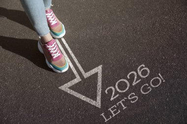 Yeni yıl 2026. Kadın okla birlikte cümleye doğru yürüyor. Hadi gidelim! Ve asfalt üzerinde sayılar, yakın plan