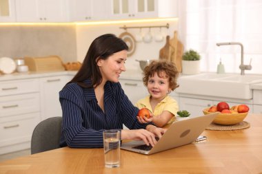 Gülümseyen anne laptopla çalışırken oğlu ev ofisinin ahşap masasında ona elma veriyor.