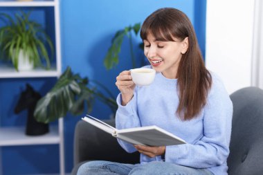 Evdeki kanepede kahve molası sırasında kitap okuyan gülümseyen kadın. Metin için boşluk
