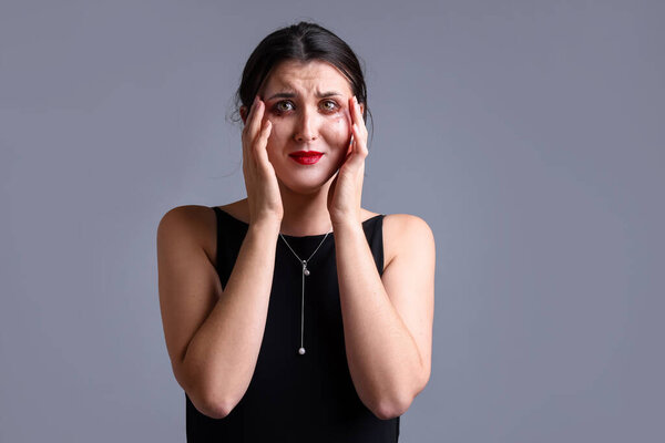 Woman with smeared mascara in dress crying on grey background