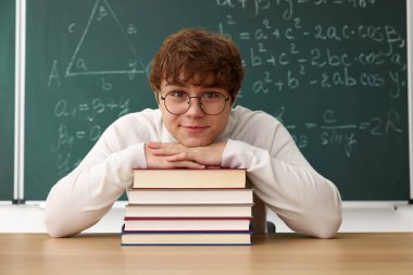 Okula dönelim. Ahşap masada kitap ve kırtasiye malzemesi olan öğrenci içeride matematik ödevi olan karatahtaya karşı.