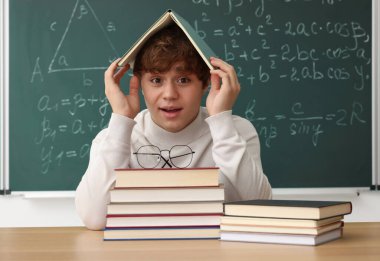 Okula dönelim. Ahşap masada kitap ve kırtasiye malzemesi olan öğrenci içeride matematik ödevi olan karatahtaya karşı.