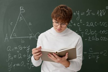 Okula dönelim. İçerde matematik ödevi olan karatahtanın yanında kitap okuyan öğrenci.