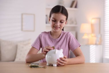 Gülümseyen küçük kız kapalı bir masadaki domuz kumbarasına para koyuyor. Cep harçlığı ve sorumluluk