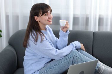 Kahve molası. Evdeki kanepede oturan elinde kupa ve dizüstü bilgisayarla gülümseyen kadın.