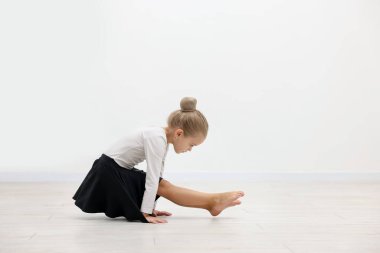 Smiling little girl doing gymnastic exercise on floor near white wall indoors. Space for text