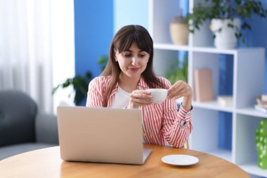 Güzel bir kadın ahşap masada laptopuyla kahve molası veriyor.