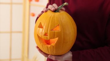 Cadılar Bayramı kutlaması. Kapalı mekanda oyulmuş ürkütücü yüzlü balkabağı tutan adam, yakın plan. Kamera hareket ediyor