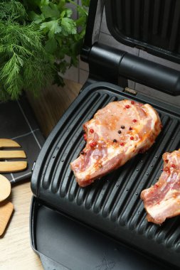 Elektrikli ızgara, pişmemiş biftekler, çatal bıçak ve otlar ahşap masada, yakın plan.