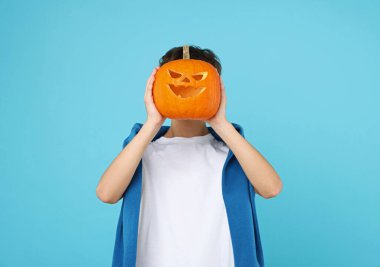 Cadılar Bayramı kutlaması. Açık mavi arka planda oyulmuş ürkütücü yüzlü balkabağı tutan çocuk.