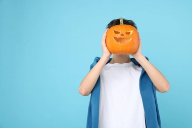 Halloween celebration. Boy holding pumpkin with carved spooky face on light blue background, space for text