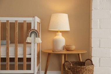 İçerideki bej duvarın yanında beşik, masa, lamba ve dekor. Şık bir bebek odası.