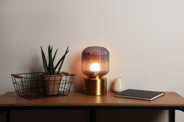 Şık lamba, vazo, çiçek ve kitap beyaz duvarın yanındaki ahşap masada.