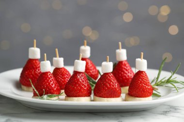 Noel yemeği. Marşmelovdan, çilekten, muzdan ve biberiyeden yapılmış Noel Baba şapkaları arka plandaki beyaz mermer masada bulanık ışıklarla, yakın plan. Bokeh etkisi