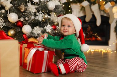 Noel Baba 'nın elf kıyafetleri içinde şirin bir bebek. Evde Noel ağacının yanında hediye kutusu tutuyor.