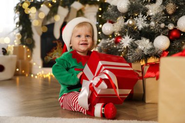 Noel Baba 'nın elf kıyafetleri içinde şirin bir bebek. Evde Noel ağacının yanında hediye kutusu tutuyor.
