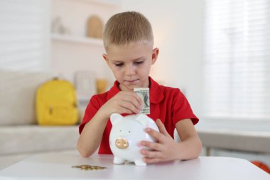 Küçük çocuk, içerideki beyaz masadaki kumbaraya cep harçlığı koyuyor.