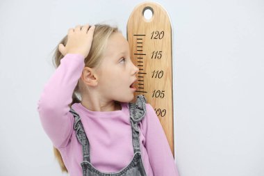 Şaşırmış küçük kız çizelgesini açık gri duvarın yanında tutuyor.