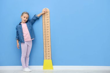 Açık mavi duvarın yanında, boyunu çizelgeyle ölçen küçük bir kız.