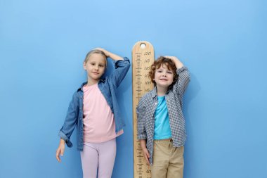 Küçük bir çocuk ve kız açık mavi duvarın yanında boylarını grafikle ölçüyorlar.