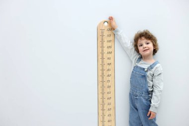 Boyunu açık gri duvarın yanında çizelgeyle ölçen küçük bir çocuk.