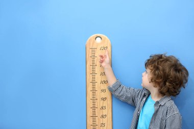 Küçük çocuk, açık mavi duvarın yanında çizelgesiyle boyunu ölçüyor.