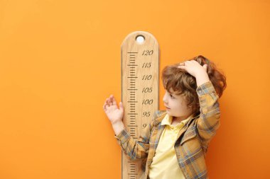 Küçük çocuk boyunu turuncu duvarın yanında çizelgeyle ölçüyor.