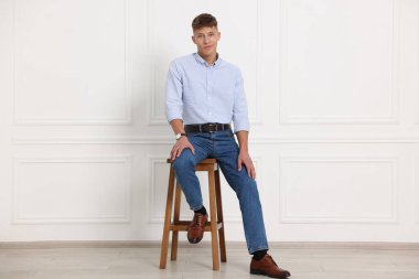 Beyaz duvarın yanındaki taburede şık kotlu yakışıklı genç adam.