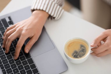 Kahve molası. Elinde bir bardak aromalı içki olan kadın ofisteki laptopta çalışıyor, yakın plan.