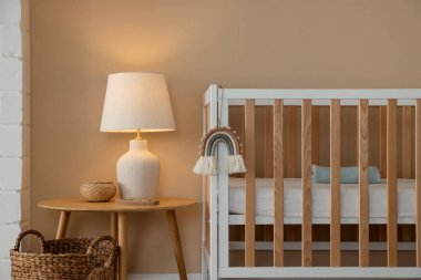 İçerideki bej duvarın yanında beşik, masa, lamba ve dekor. Şık bir bebek odası.