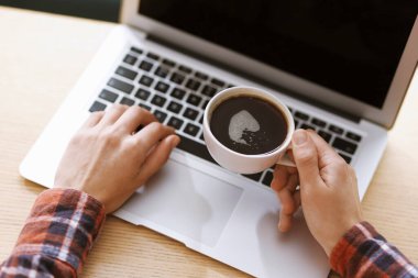 Kahve molası. Elinde bir bardak aromalı içki olan kadın ofisteki laptopta çalışıyor, yakın plan.