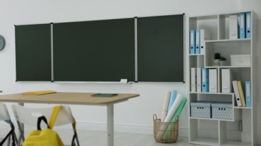 Sınıfta yeşil karatahta, sırt çantaları ve şık mobilyalar. Kamera sola hareket ediyor