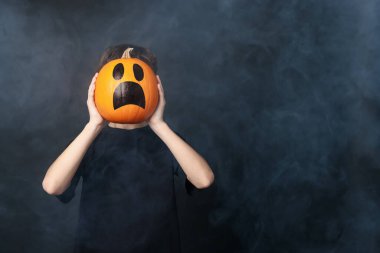 Cadılar Bayramı kutlaması. Siyah zemin üzerinde çizilmiş ürkütücü yüzü olan balkabağı tutan çocuk dumanlı, mesaj için alan