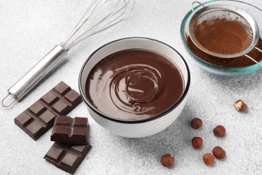 Erimiş çikolata kasede, parçalar, fındıklar, kakao tozu ve çırpı açık gri masada, yakın plan.