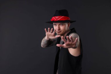 Piercingli, dövmeli ve koyu gri arkaplanda poz veren şık elbiseli genç bir adam.