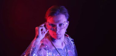 Piercingli, dövmeli ve siyah arka planda neon ışıklarla poz veren genç bir adam.