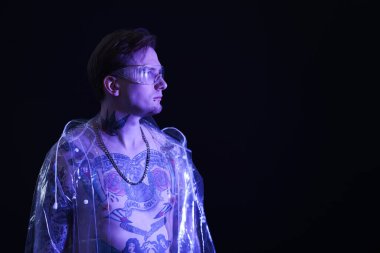 Piercingli, dövmeli ve şık elbiseli genç adam siyah arka planda neon ışıklarıyla poz veriyor.