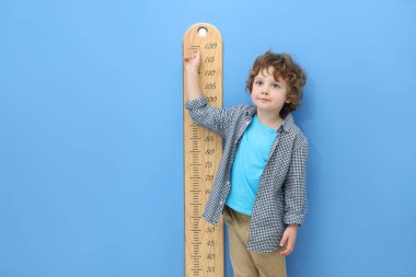 Küçük çocuk, açık mavi duvarın yanında çizelgesiyle boyunu ölçüyor.