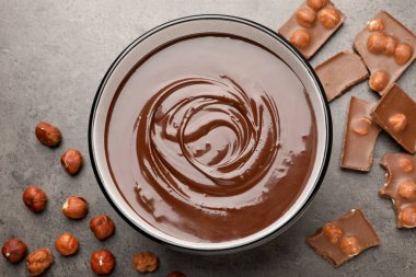 Erimiş çikolata kasede, parçalar ve fındıklar gri masada, düz yerde