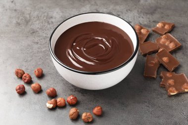 Erimiş çikolata kasede, parçalar ve fındıklar gri masada, yakın plan.