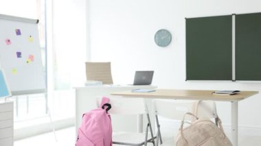 Sınıfta yeşil karatahta, sırt çantaları ve şık mobilyalar. Kamera sağa hareket ediyor