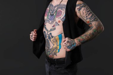 Dövmeli ve şık elbiseli genç adam koyu gri arka planda poz veriyor, yakın plan.