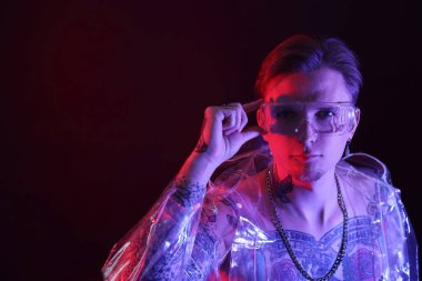 Piercingli, dövmeli ve şık elbiseli genç adam siyah arka planda neon ışıklarıyla poz veriyor.