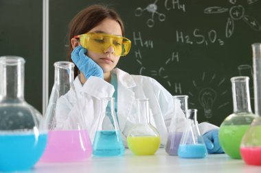 Okula dönelim. Masada elinde sıvı şişeleriyle oturan üzgün bir kız var. Kapalı alanda kimyasal formüllü karatahtaya karşı.