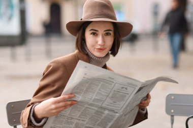 Şapkalı ve takım elbiseli zarif bir kadın portresi ve açık hava kafesinde gazete.