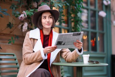 Şapkalı ve ceketli güzel bir kadın açık kafedeki masada gazete okuyor.