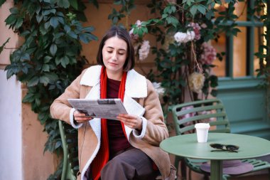 Paltolu güzel bir kadın açık kafede gazete okuyor.