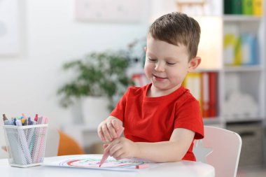 Smiling little boy with felt pen drawing at white table indoors